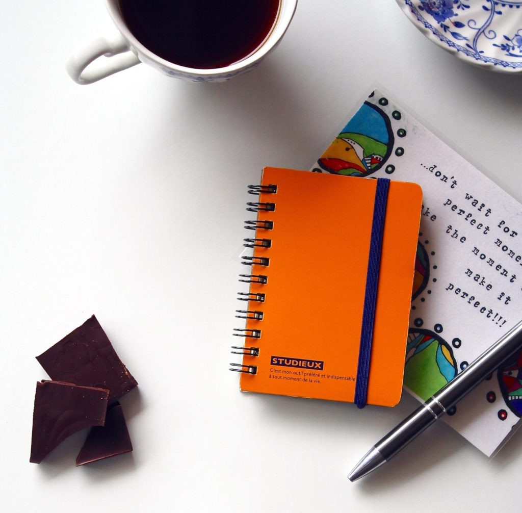 The little things are what can matter the most imiage may contain: an orange notebook, grey pen, chocolate, coffee in a cup, a card, and the corner of a saucer on a table signifying needs of residents at Hannah House